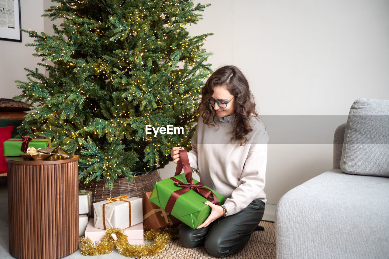 A cute girl with glasses is sitting on the carpet near the christmas tree and unpacking gifts