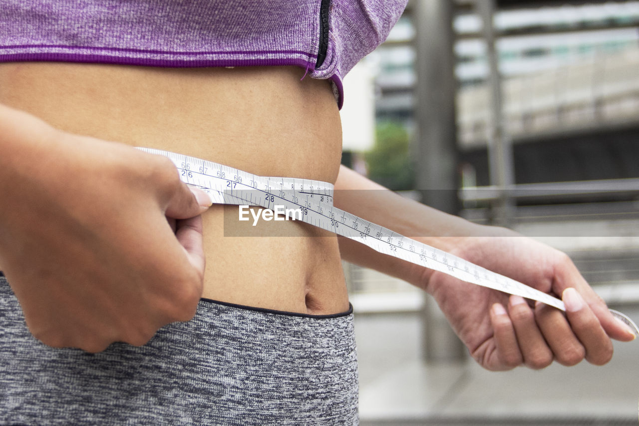 Midsection of woman measuring waist while standing outdoors