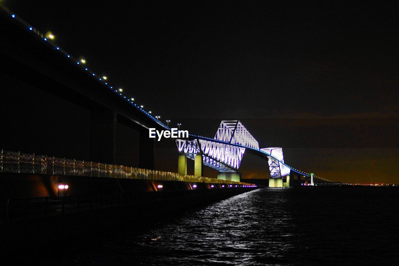 LOW ANGLE VIEW OF ILLUMINATED BRIDGE OVER RIVER AT NIGHT