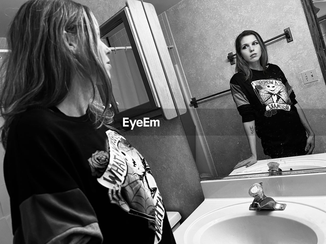 Black and white portrait of young woman standing in bathroom looking in a mirror.