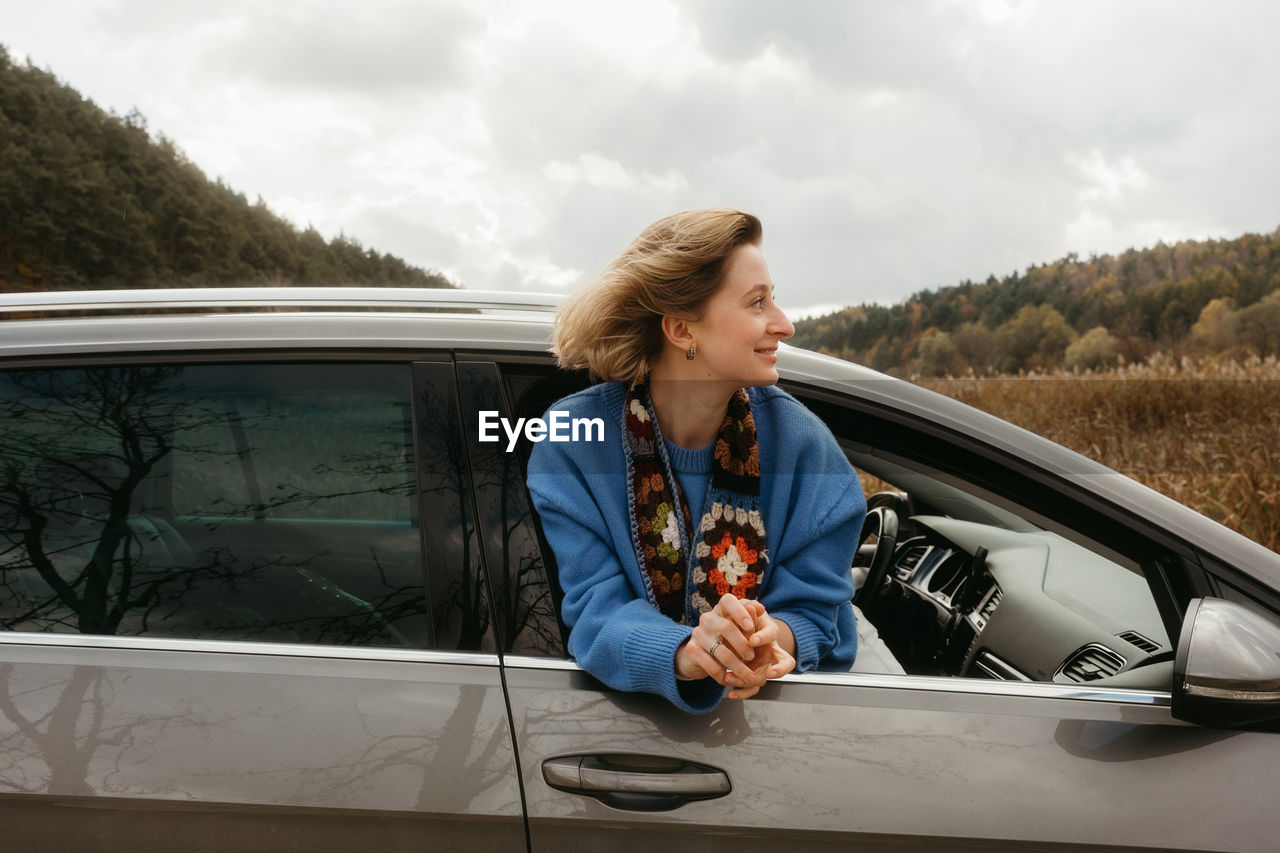 A woman leans out of the car window during road trip in autumn, the wind blows her hair