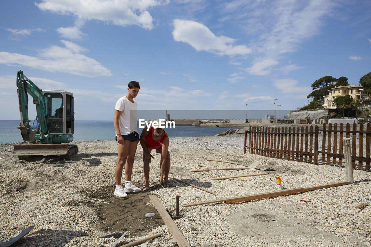 REAR VIEW OF MEN STANDING ON SHORE AGAINST SKY