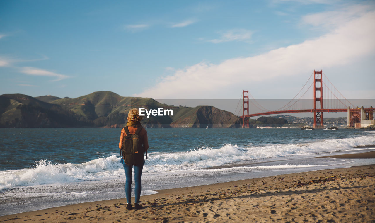 Rear view of woman standing on shore with golden gate bridge in background