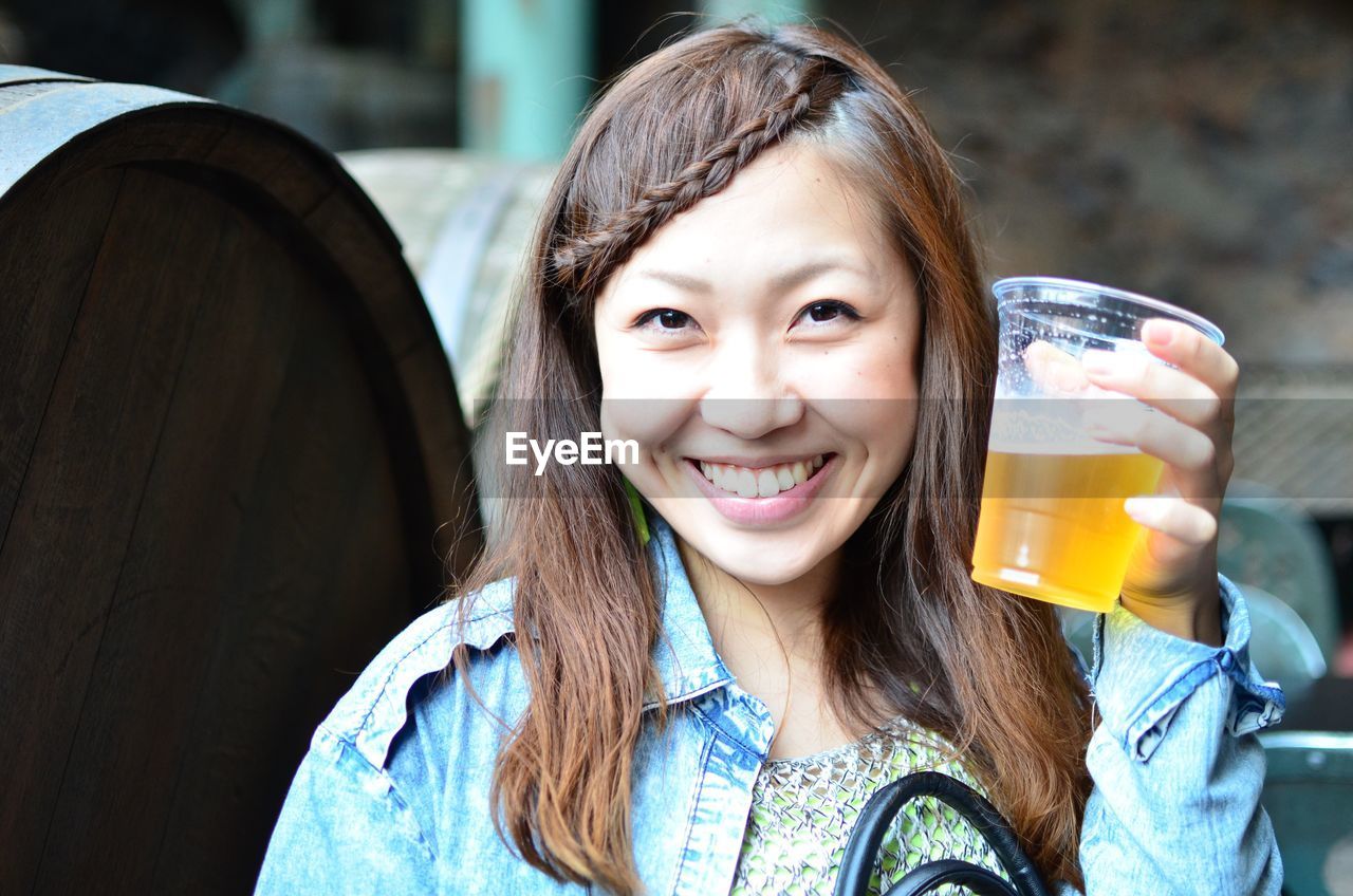 Smiling young woman holding beer glass against barrels on sunny day
