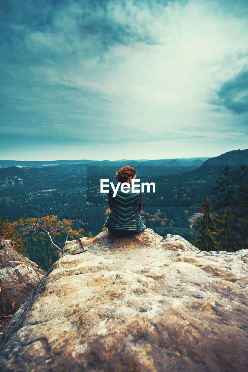 REAR VIEW OF YOUNG WOMAN SITTING ON MOUNTAIN