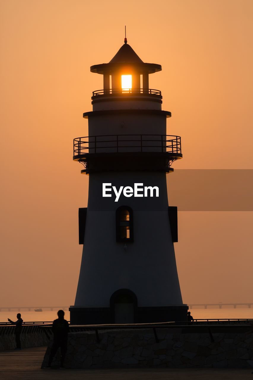 Silhouette building by lighthouse against sky during sunset
