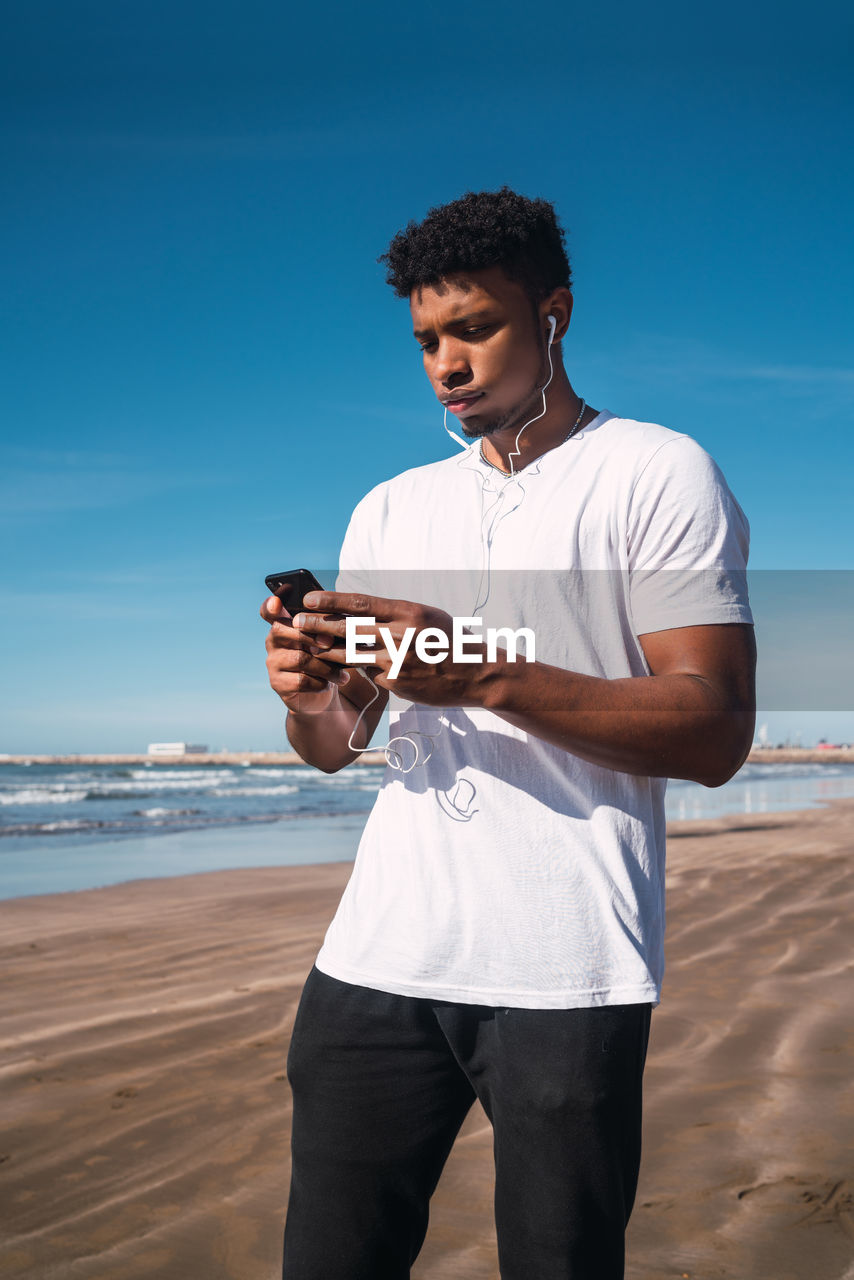 Young man using mobile phone while standing at beach against clear blue sky