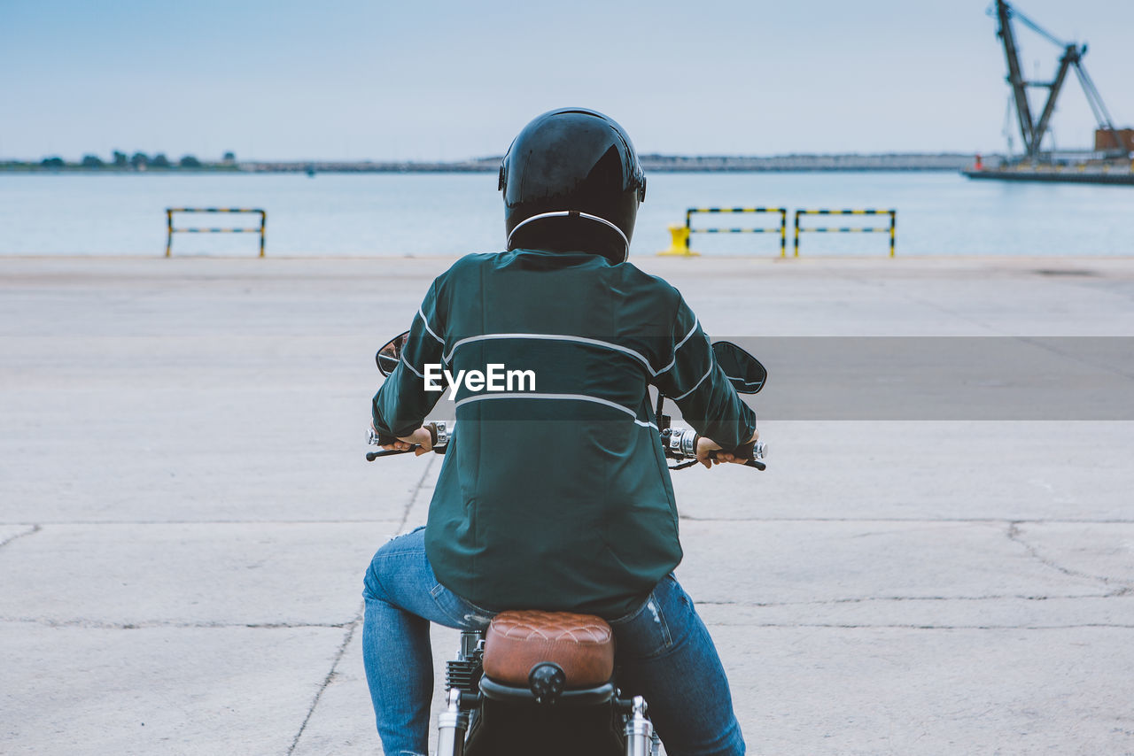 Back view of unrecognizable male biker in casual outfit and protective helmet sitting on motorcycle on embankment near sea at construction site