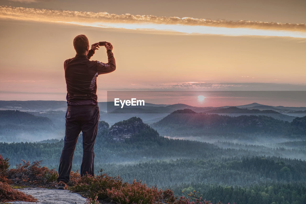 Man holding camera at eyes on mountain and watching sunset.