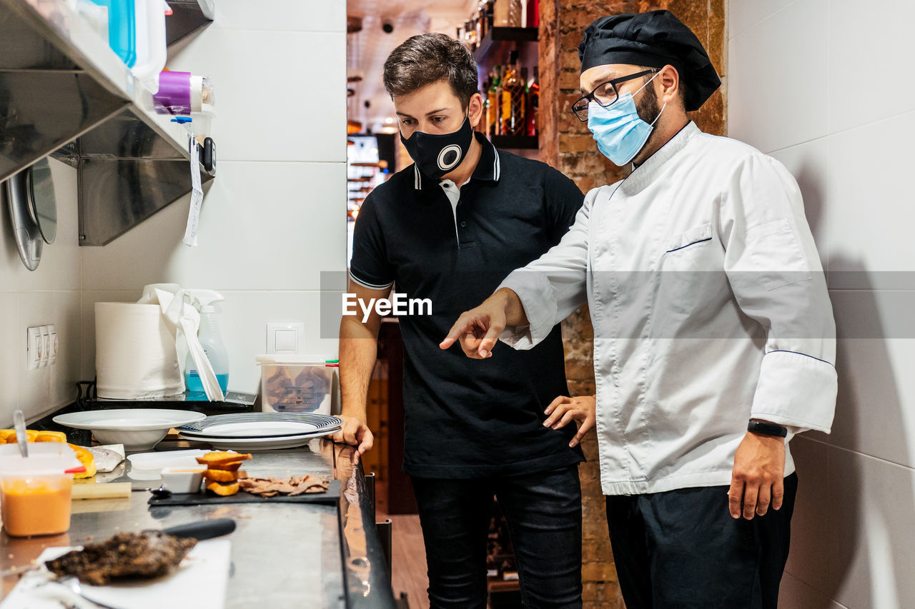 Male cook in medical mask talking to waiter and pointing at prepared delicious dish ready for serving in cafe