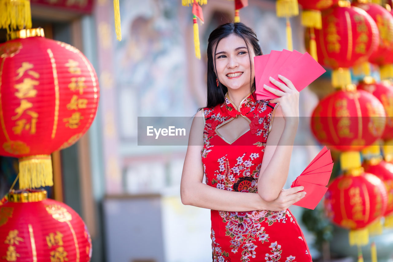 PORTRAIT OF SMILING YOUNG WOMAN IN TRADITIONAL CLOTHING STANDING AGAINST RED WALL