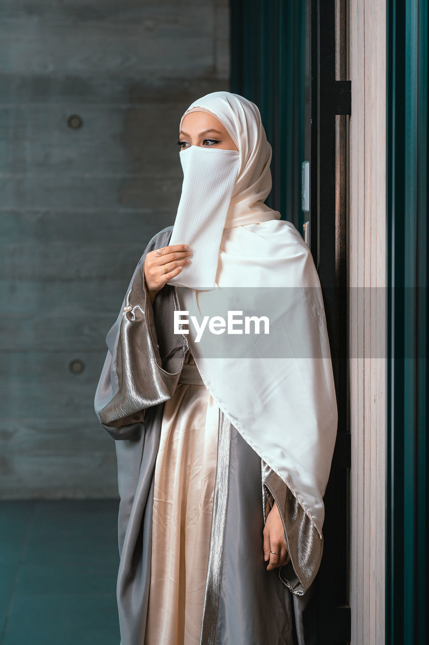 Front view portrait of veiled woman with white niqab and hijab standing against the wall