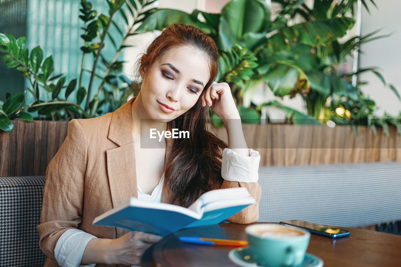 Young brunette asian girl student reading book with coffee on the table at cafe