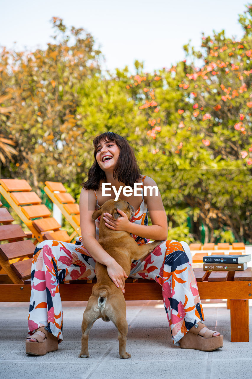 Young woman with dog sitting outdoors next to a pool, in summertime.