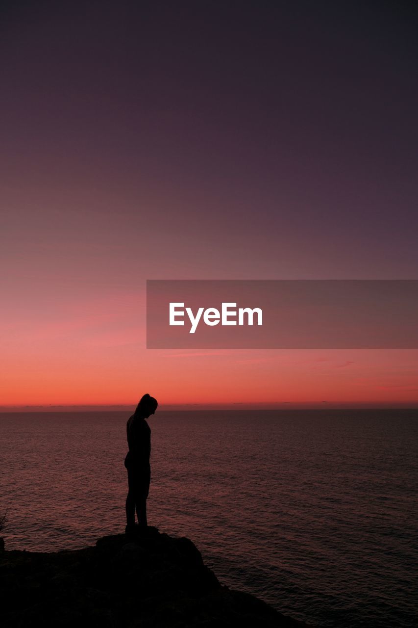 Side view of silhouette woman standing by sea against dramatic sky during sunset