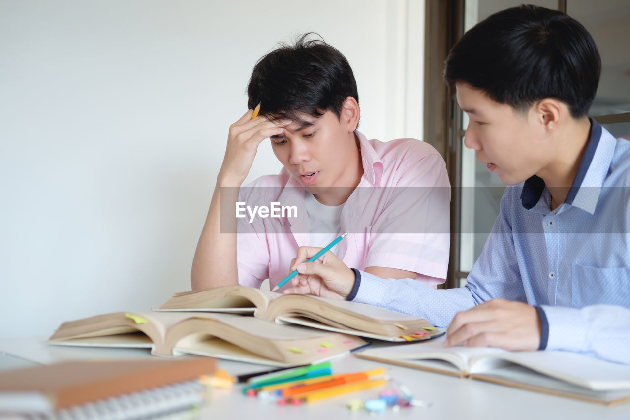 Young man assisting friend in studying at table in classroom