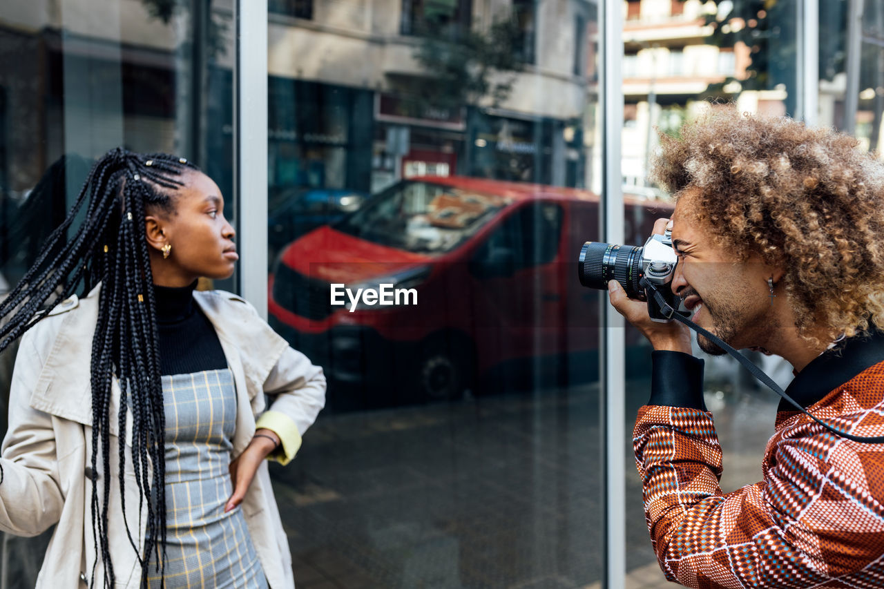 Creative male photographer with camera shooting photo of stylish young african american female model with afro pigtails near glass wall of urban building