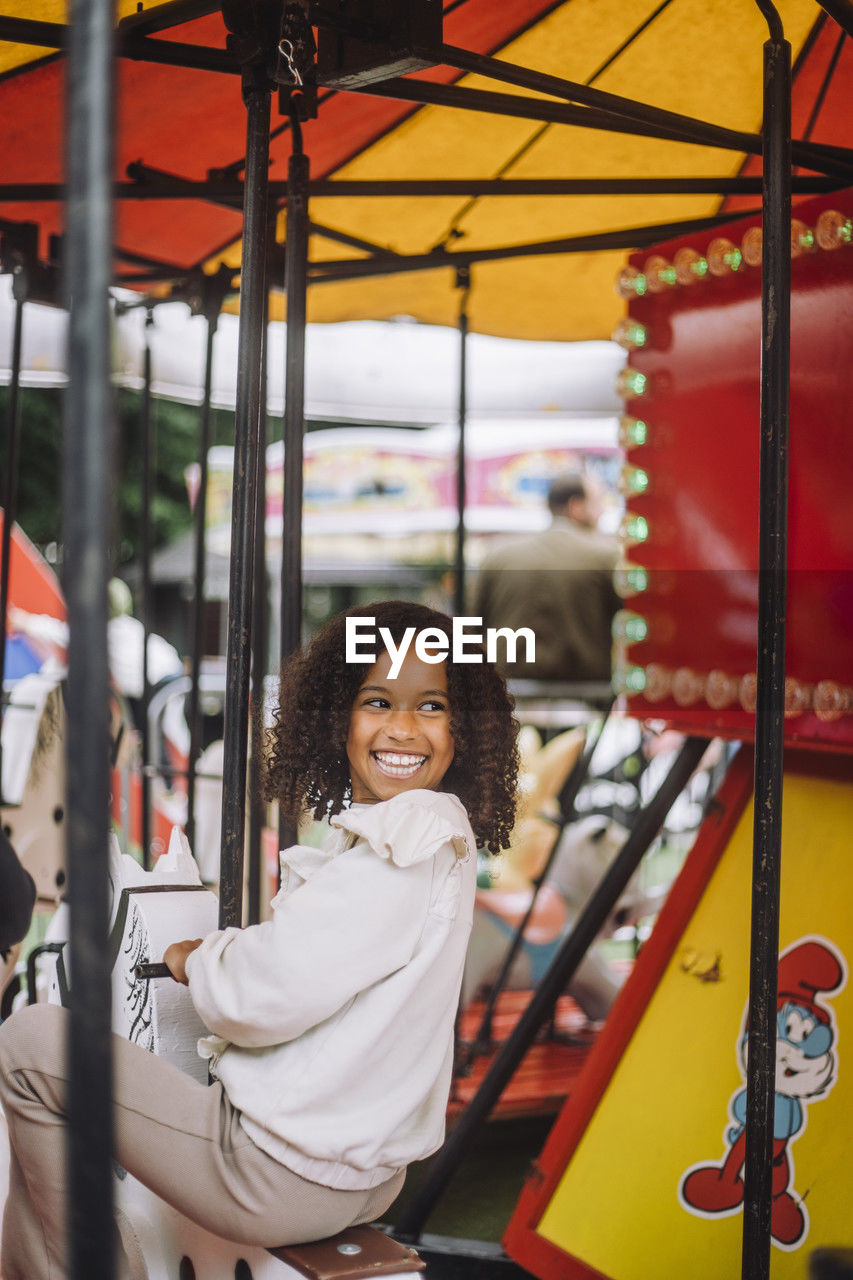 Happy girl looking over shoulder while having fun on carousel at amusement park