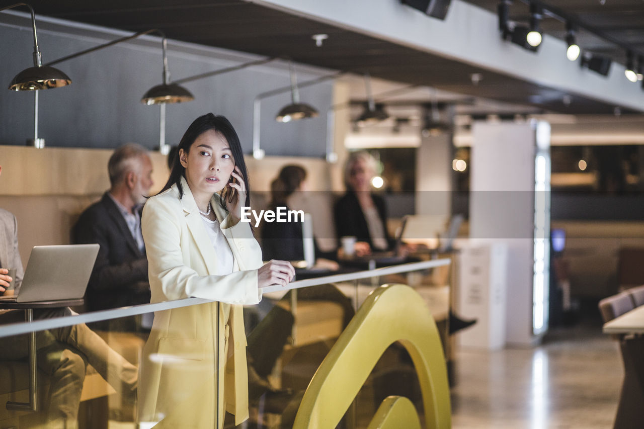 Female entrepreneur talking on mobile phone while standing by railing with colleagues in background at office