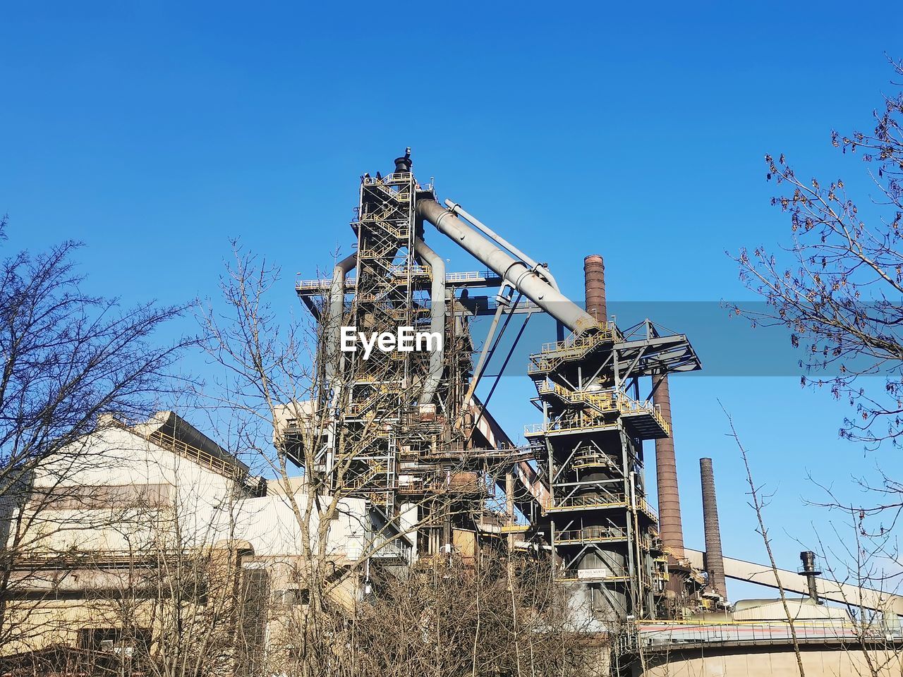 sky, architecture, blue, industry, tree, nature, bare tree, built structure, clear sky, plant, no people, day, low angle view, sunny, power generation, outdoors, machinery, building exterior, sunlight