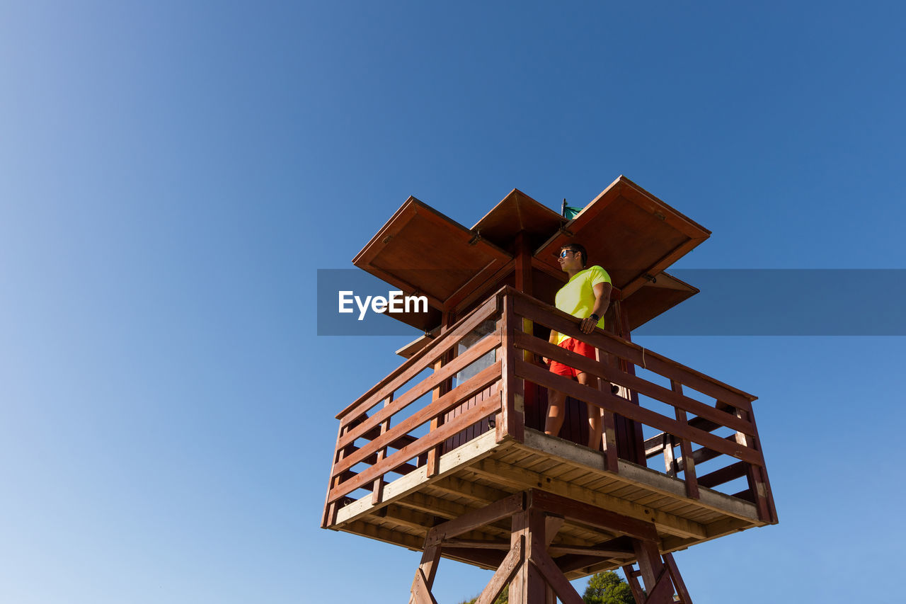 From below male lifeguard controlling safety on sea from wooden rescue tower