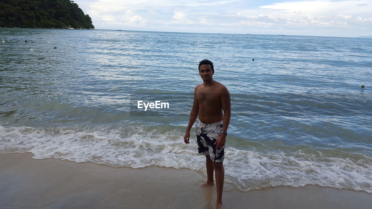 FULL LENGTH OF SHIRTLESS MAN STANDING AT BEACH