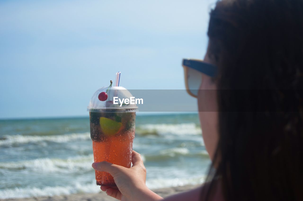 Close-up of woman holding drink at beach against sky