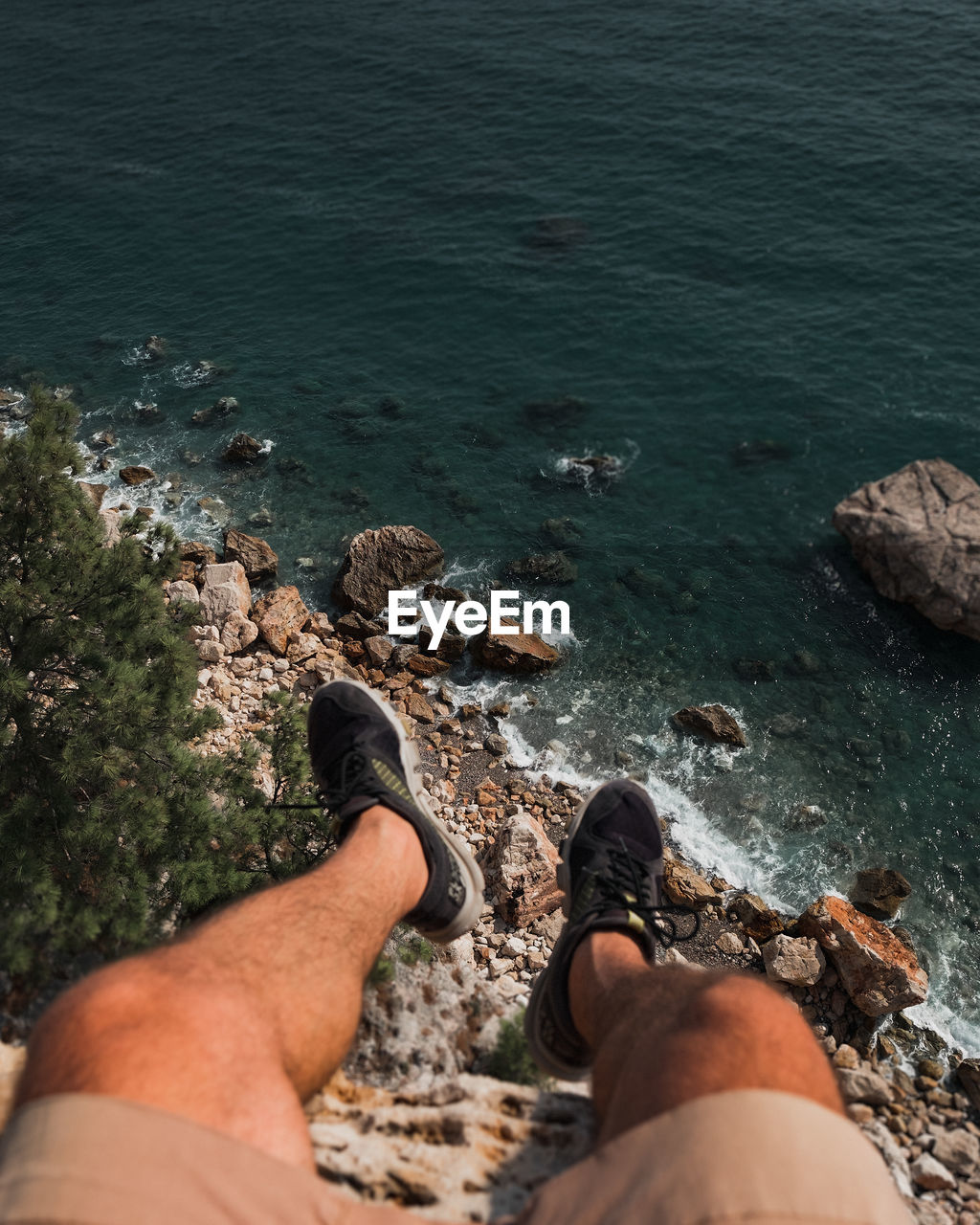 Legs of man are hanging from edge of cliff over the sea and rocks.