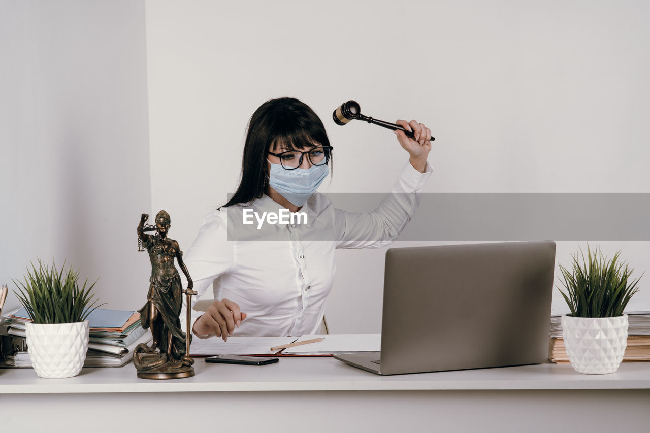 Young frustrated lawyer wearing mask hitting laptop by hammer at office