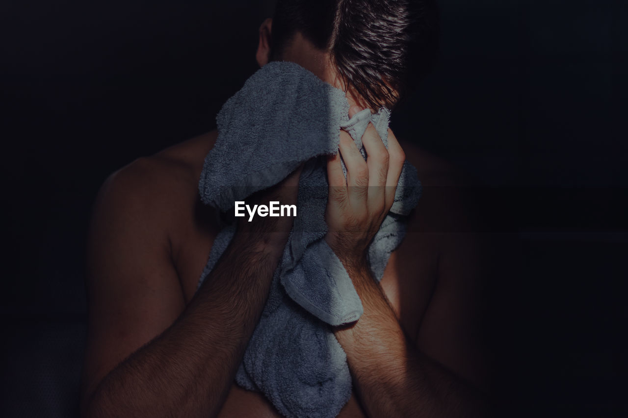 Shirtless man covering face with towel while standing in darkroom