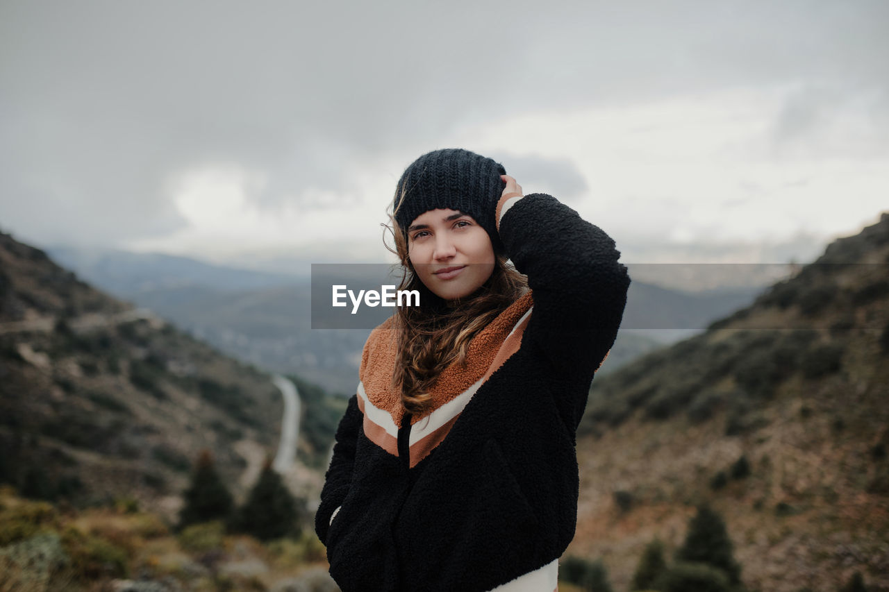 Smiling young female putting on warm hat and looking at camera while standing on rough highlands on cloudy gloomy day