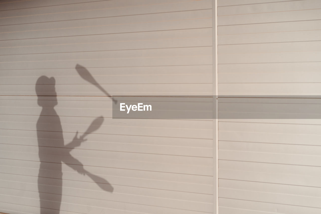 Side view shadow of anonymous talented male circus performer in hat juggling clubs near white wall