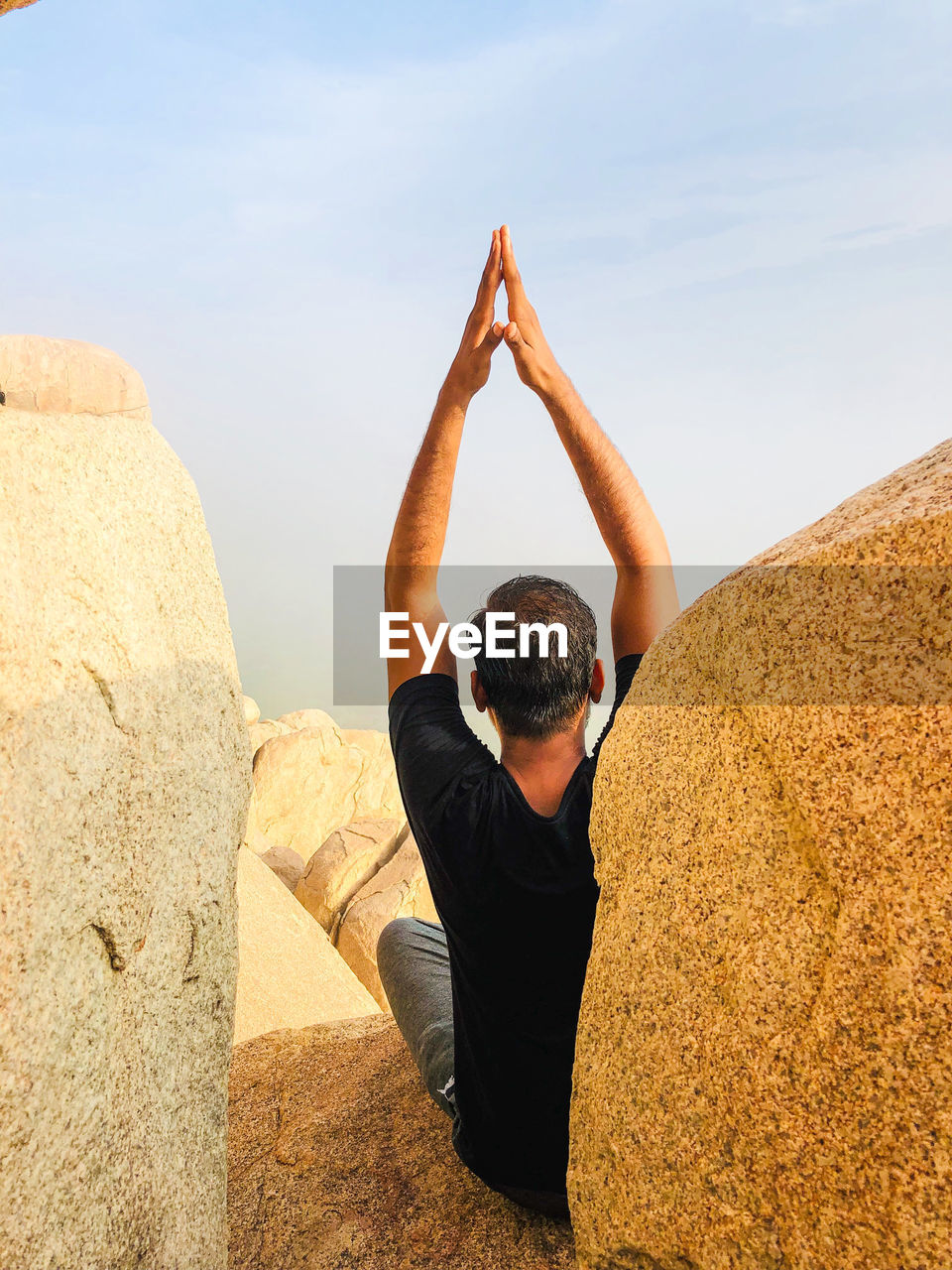 Rear view of man with arms raised meditating while sitting on rock against sky