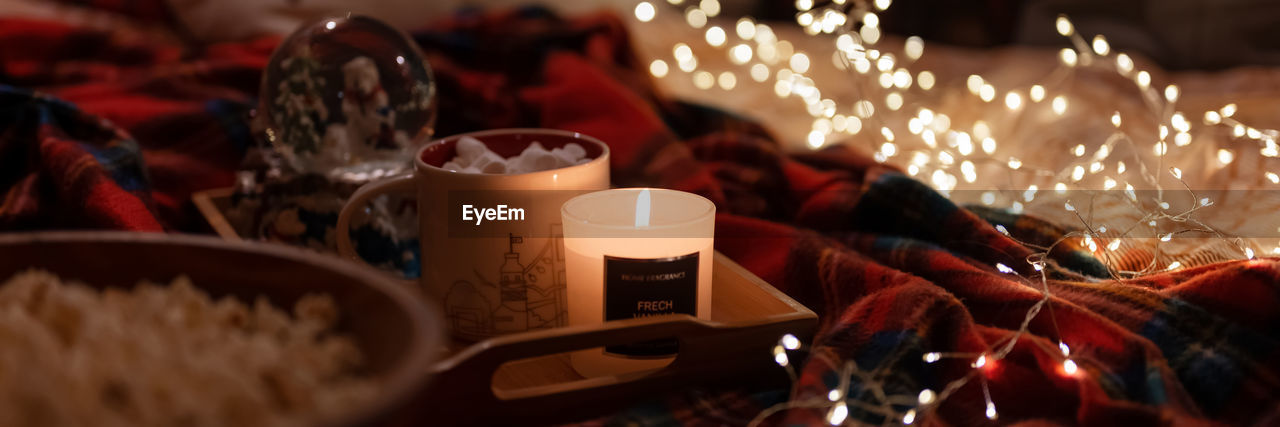 Cup of hot chocolate and marshmallows and candle on tray bed and glowing lights bokeh at home