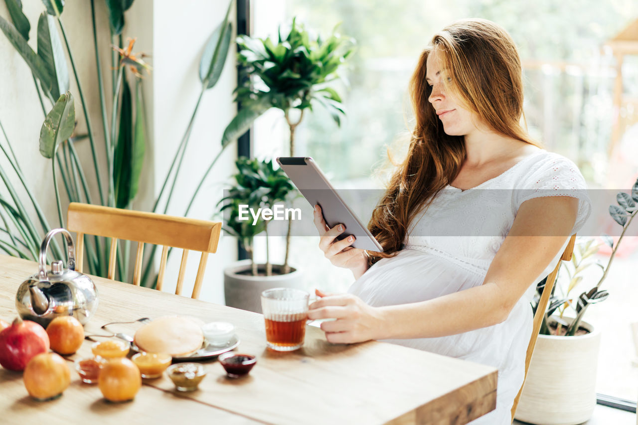 A pregnant woman is eating breakfast at the table and looking into a tablet. communication