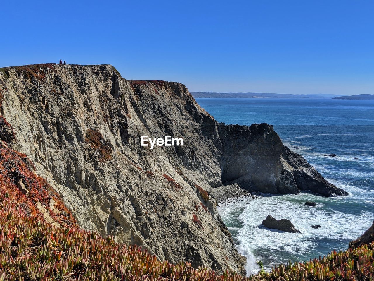 People so small in contrast to ocean cliffs against blue background.