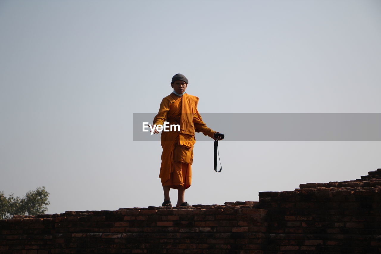 Portrait of monk standing on brick wall against clear sky