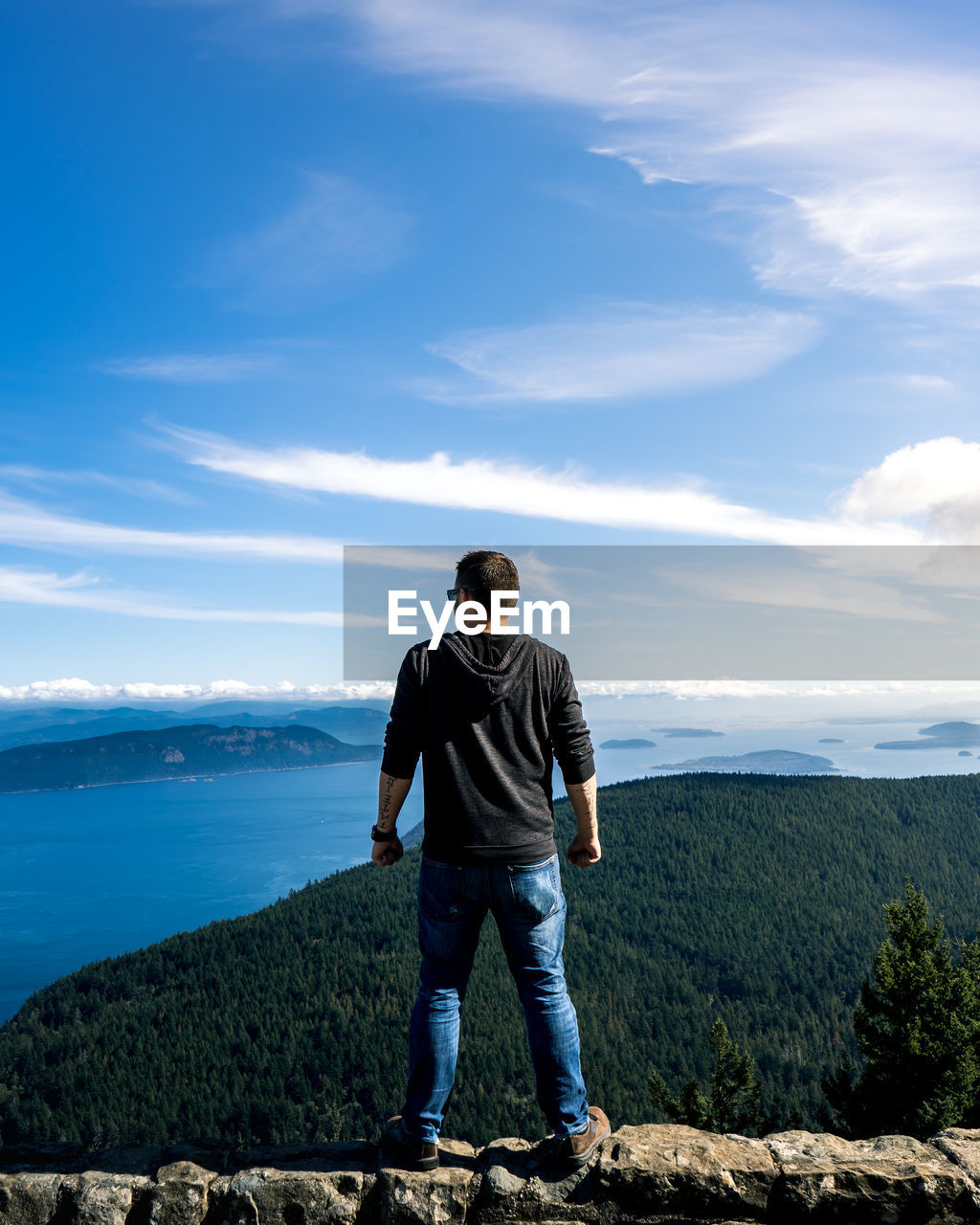 Rear view of man standing on railing with sea in background against blue sky