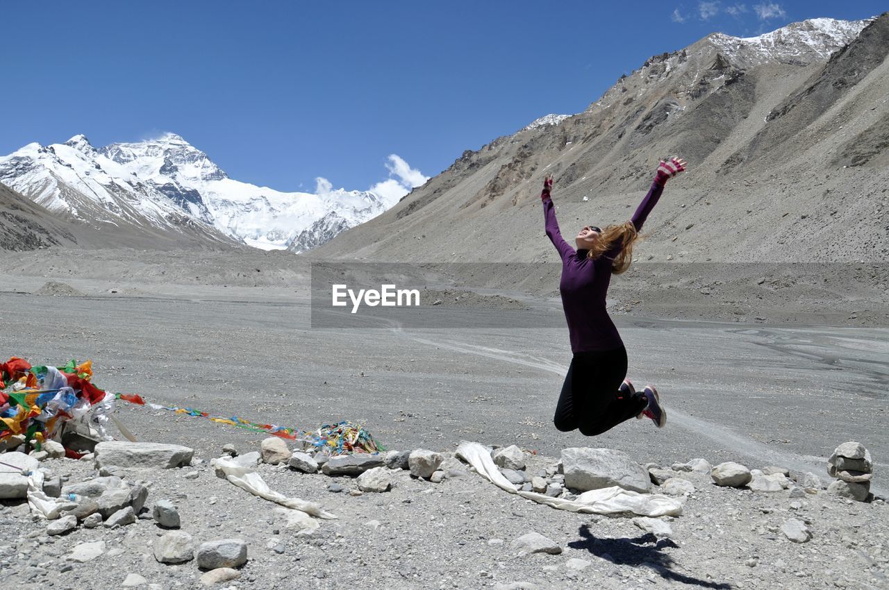 Full length of woman jumping in mid-air over rocks against snowcapped mountains