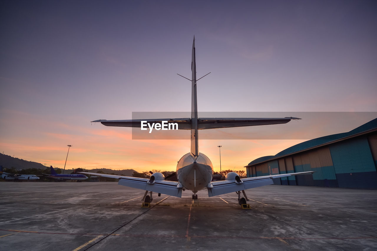 REAR VIEW OF AIRPLANE AT AIRPORT RUNWAY AGAINST SKY DURING SUNSET