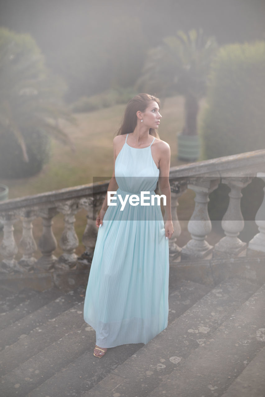 women, adult, one person, fashion, wedding dress, young adult, dress, female, bridal clothing, full length, clothing, bride, gown, nature, standing, wedding, elegance, celebration, formal wear, event, newlywed, emotion, blue, hairstyle, looking, white, outdoors, glamour, bridal party dress, lifestyles, happiness, contemplation, portrait, day, beauty in nature, brown hair, sky, life events, evening gown, architecture, blond hair, looking away, strapless, copy space, water, person, sunlight, tranquility, smiling