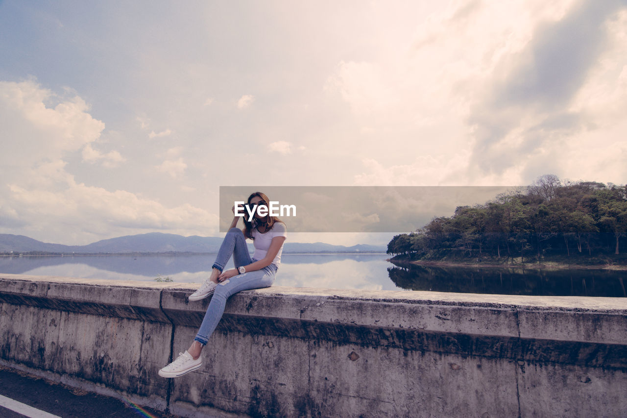 Portrait of woman photographing while sitting against lake and sky