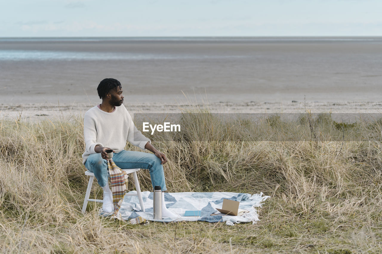 Young man drinking tea while sitting on stool amidst dried plants at beach