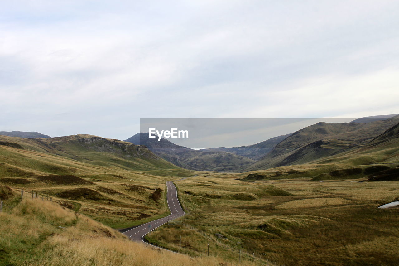 Road amidst grassy field against cloudy sky at cairngorms national park