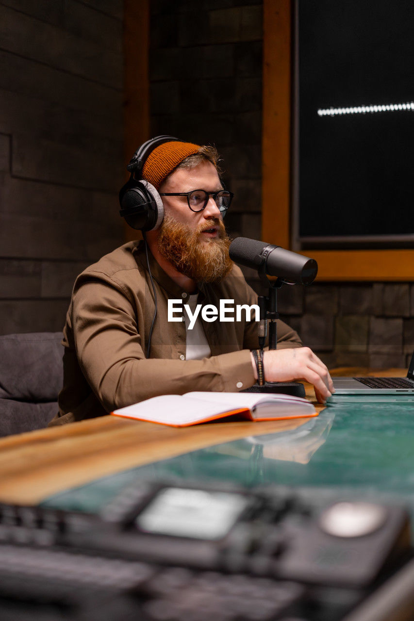 A man with a beard and headphones is seated at a table in front of a microphone