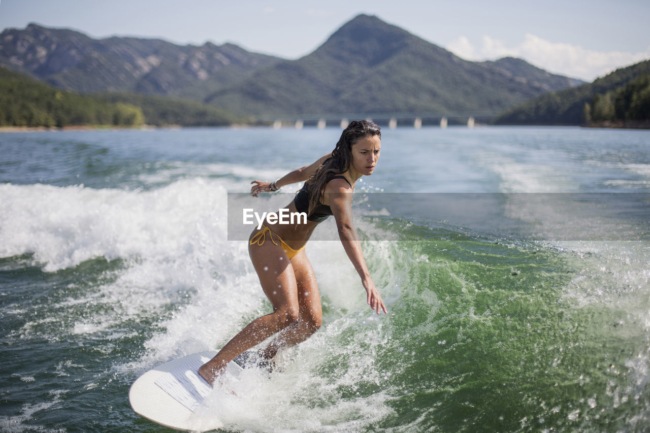 FULL LENGTH OF WOMAN SURFING ON SEA AGAINST MOUNTAINS
