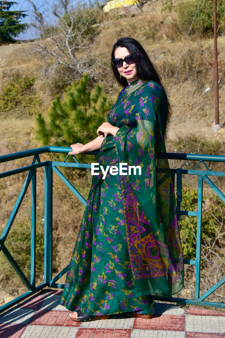 one person, fashion, women, adult, clothing, young adult, dress, full length, green, portrait, spring, standing, looking at camera, photo shoot, lifestyles, nature, railing, blue, hairstyle, traditional clothing, plant, smiling, day, leisure activity, female, outdoors, front view, glasses, tree, long hair, architecture, costume