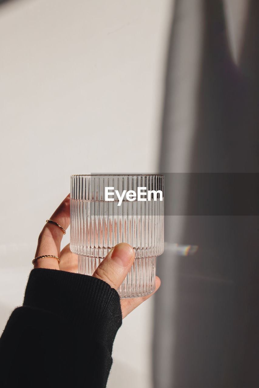 Woman's hand holds a glass of clean water over the white wall in natural sunlight