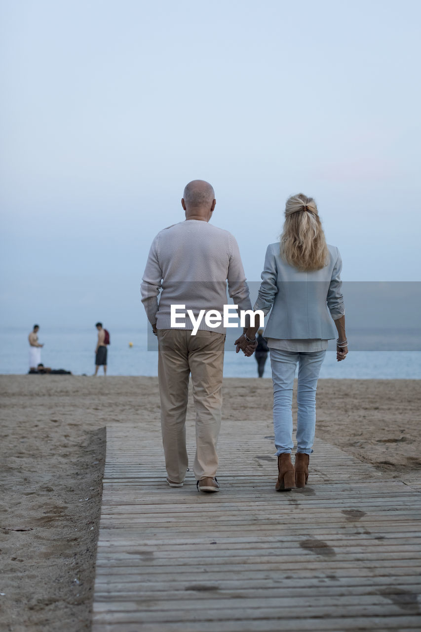 Spain, barcelona, rear view of senior couple walking hand in hand on the beach at dusk