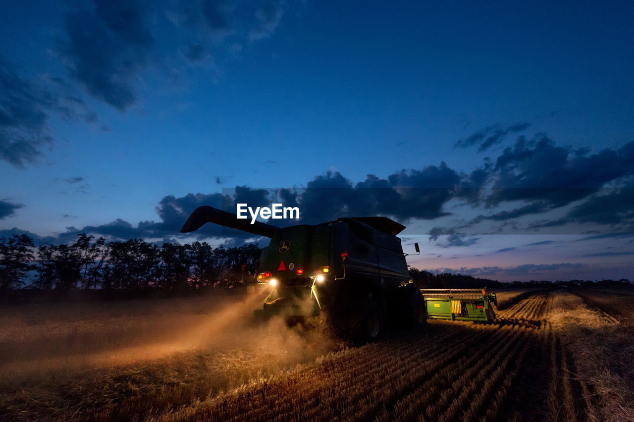 Illuminated combine harvester on agricultural field against sky at dusk